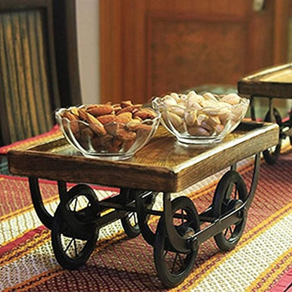 Wooden serving cart with wheels on a striped rug, containing glass bowls of snacks.